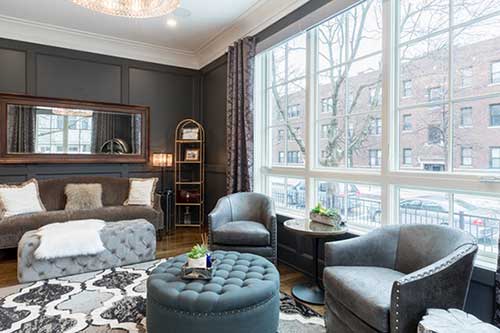 A modern living room with dark walls, large windows, gray armchairs, a tufted ottoman, sofa with pillows, patterned rug, and gold shelving unit—perfectly styled for a real estate web design photo shoot. Bright light streams in from the city street outside. - Clio Websites | Top Canadian Web Design Company