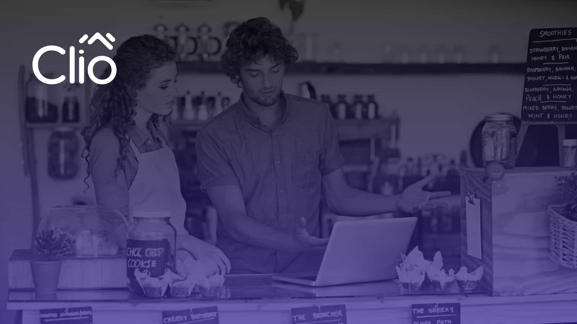 Two people stand behind a cafe counter, looking at a laptop—one gestures toward the screen while the other observes. The Clio logo appears in the top left corner. Shelves with jars and menu boards fill the background, suggesting small business website packages in use. - Clio Websites | Top Canadian Web Design Company