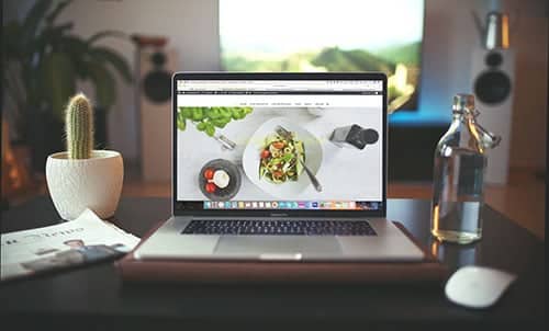 A laptop on a desk displays a food website, highlighting small business website packages. Beside it are a small potted cactus, a magazine, a computer mouse, and a clear glass bottle of water. The background shows a blurred TV and living room decor. - Clio Websites | Top Canadian Web Design Company