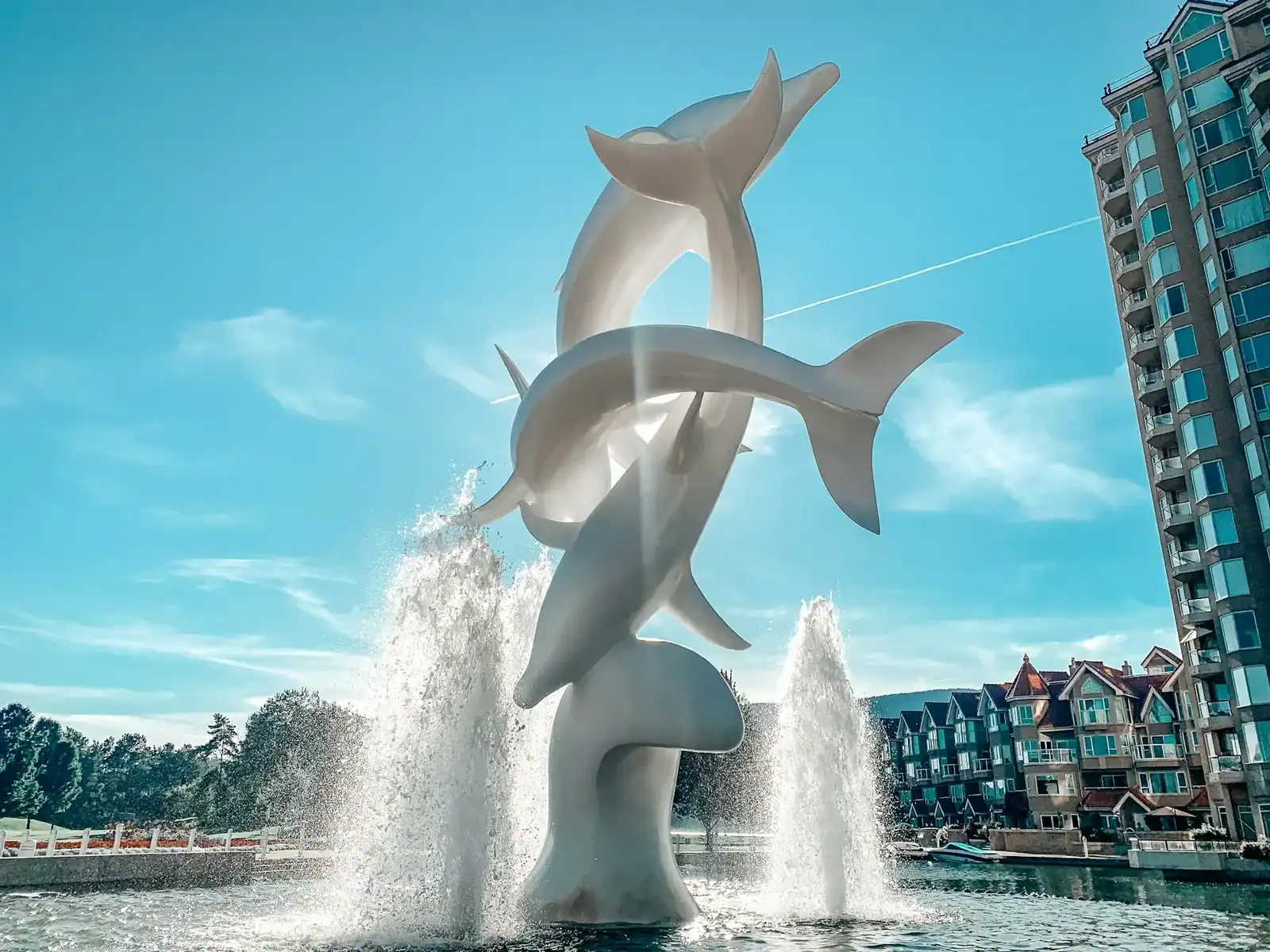 A tall, white sculpture of three dolphins leaping among water fountains, set against a blue sky with modern buildings in the background—much like the creativity and elegance you’d find in standout Kelowna web design. - Clio Websites | Top Canadian Web Design Company