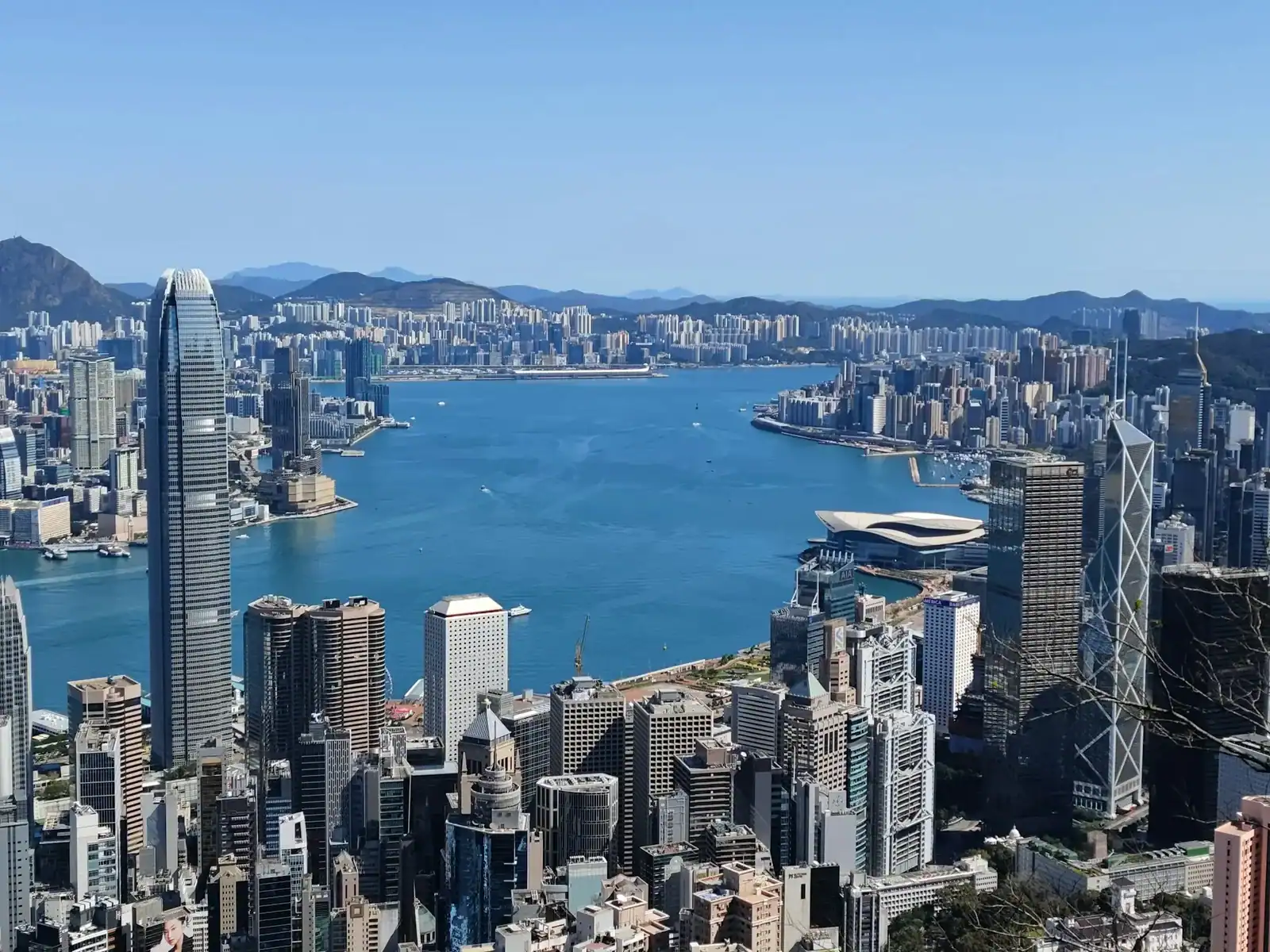 High-angle view of Hong Kong’s skyline with tall skyscrapers, Victoria Harbour in the center, and distant mountains under a clear blue sky—an inspiring scene reminiscent of the innovation and creativity you’ll find at Victoria Web Design. - Clio Websites | Top Canadian Web Design Company