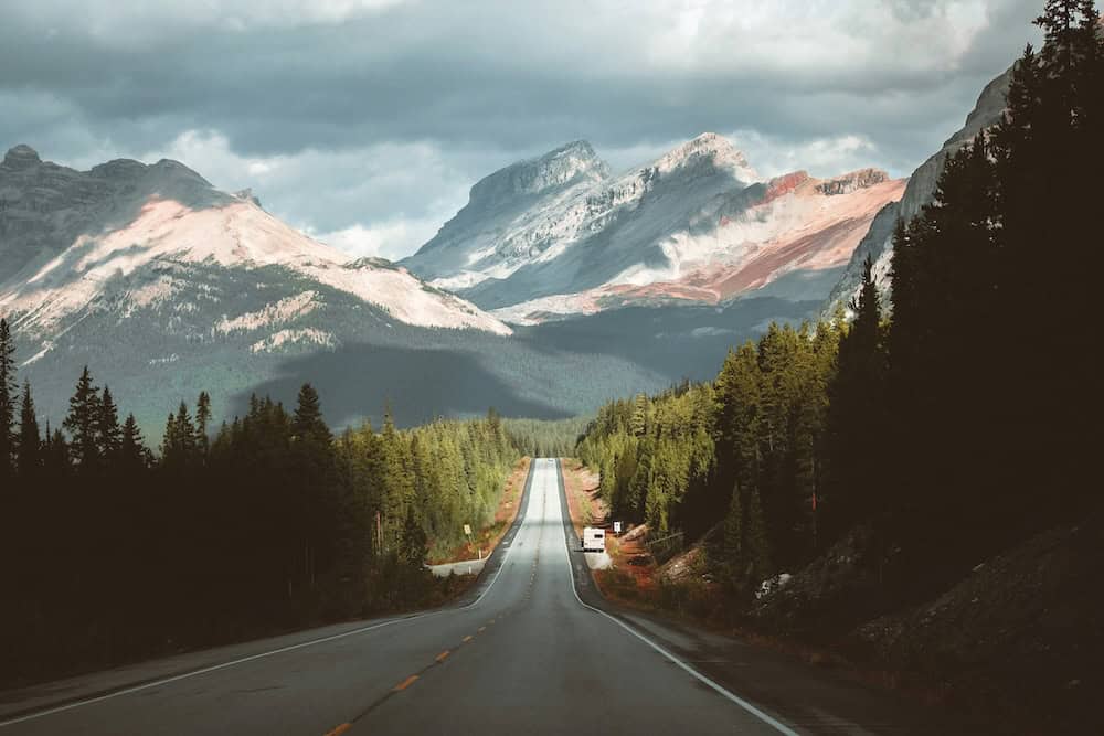 A long, empty road stretches through a forest towards distant, jagged mountains under a cloudy sky. Sunlight illuminates parts of the peaks, creating a dramatic and scenic landscape—much like the creative journey in Alberta web design. - Clio Websites | Top Canadian Web Design Company