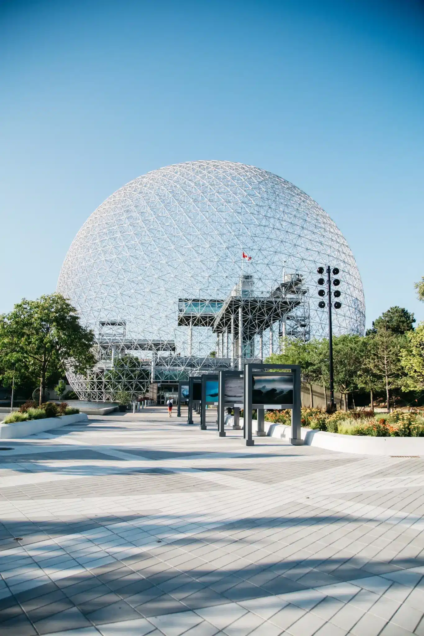 A large geodesic dome structure with a metal framework, surrounded by trees under a clear blue sky, evokes the innovative spirit of Montreal web design. The pathway leading to it is lined with informational displays and lampposts. A flag is visibly fluttering atop the structure. - Clio Websites | Top Canadian Web Design Company