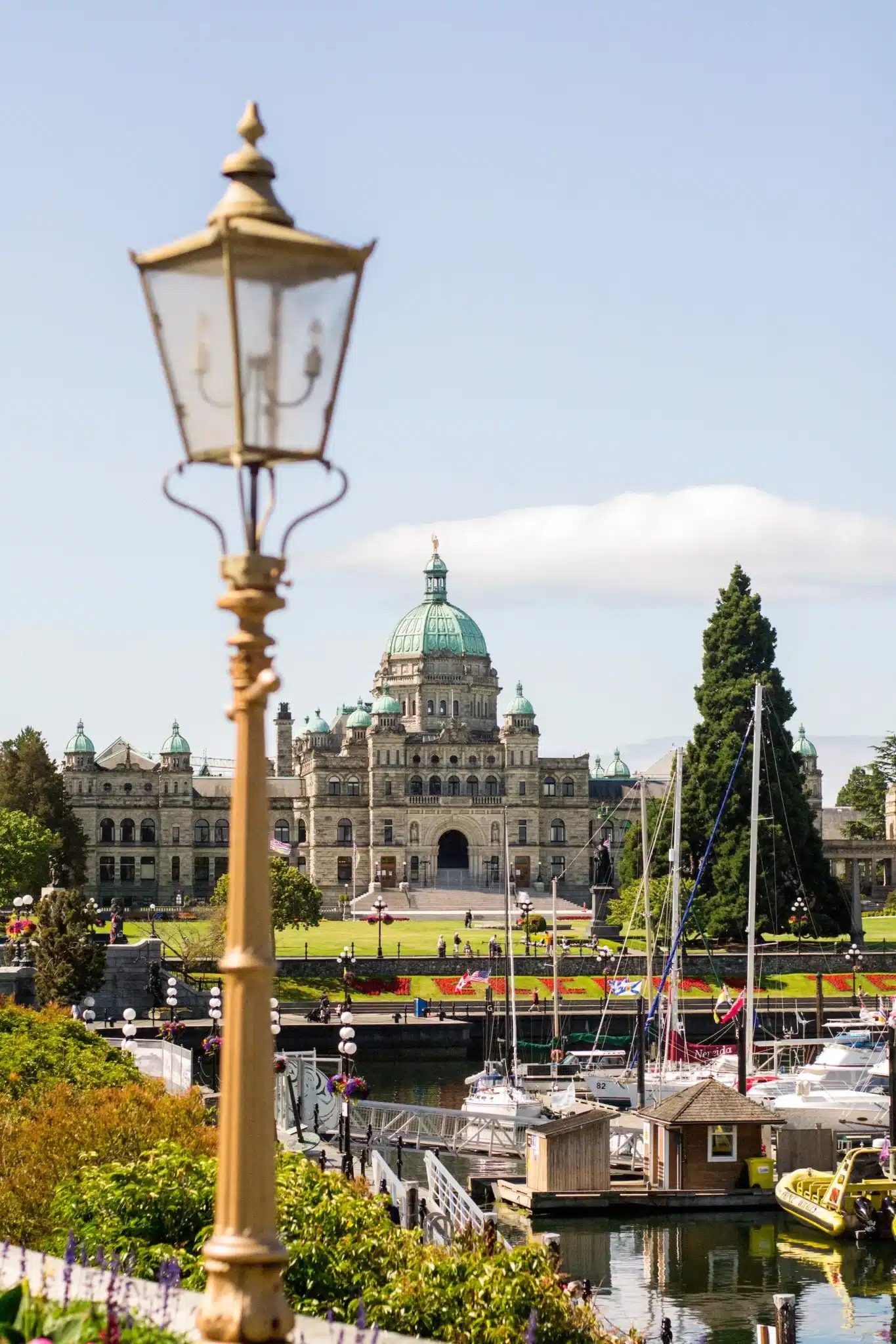 View of the British Columbia Parliament Buildings with a vintage street lamp in the foreground, reminiscent of Victoria's classic charm. Boats are docked in the harbor under a clear sky, while lush greenery and colorful flowers enhance this picturesque scene. - Clio Websites | Top Canadian Web Design Company