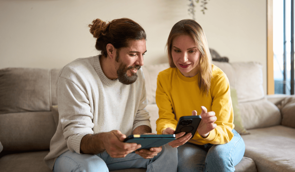 A man and woman sit on a couch in Canada, smiling as they look at a phone in her hand. The man holds a tablet, and together they explore an interesting 2026 Guide to Managed WordPress Hosting on their devices. - Clio Websites | Top Canadian Web Design Company