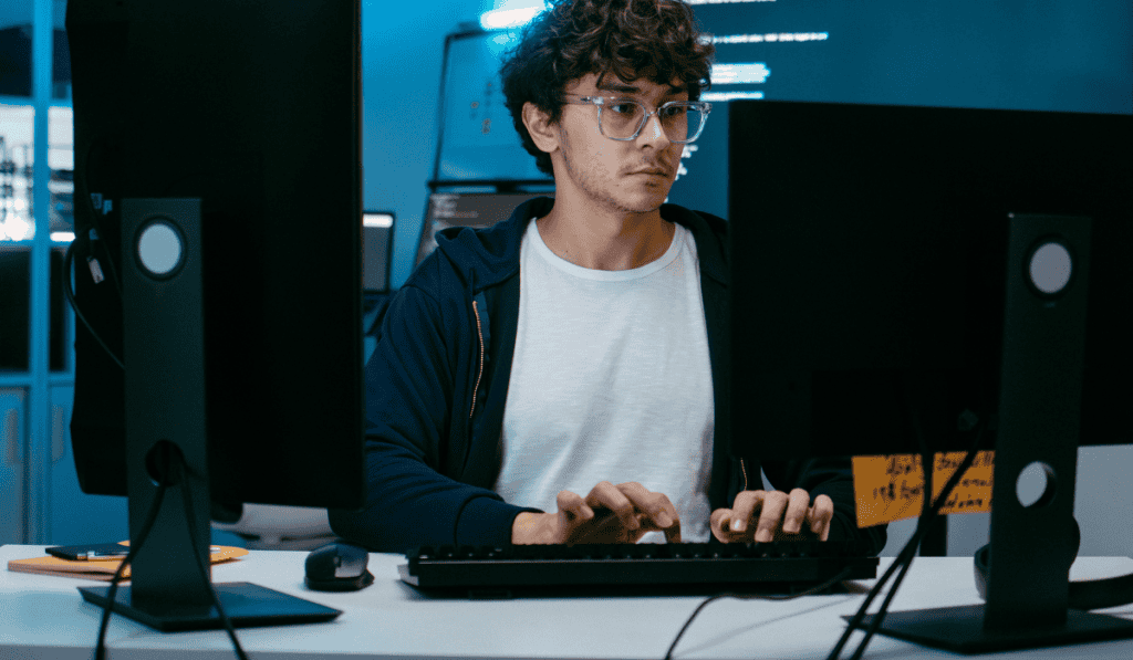 A person wearing glasses and a white t-shirt is sitting at a desk, typing on a keyboard between two computer monitors in a dimly lit room with blue lighting, focused on Managed WordPress Hosting Canada solutions. - Clio Websites | Top Canadian Web Design Company