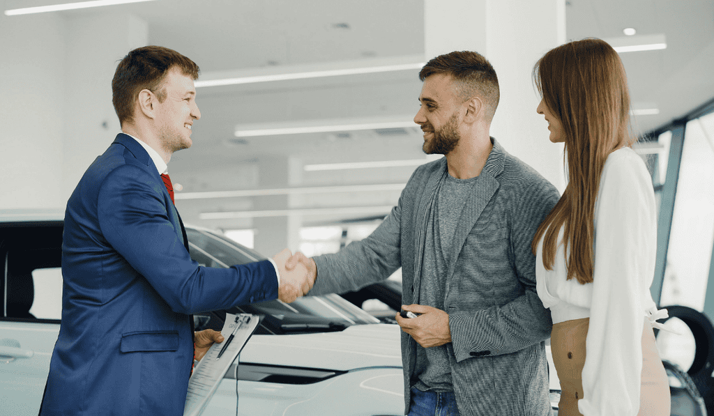 A car salesman in a blue suit shakes hands with a smiling man next to a woman in a white blouse, standing beside a white car in a brightly lit showroom that exemplifies Calgary website accessibility standards. - Clio Websites | Top Canadian Web Design Company