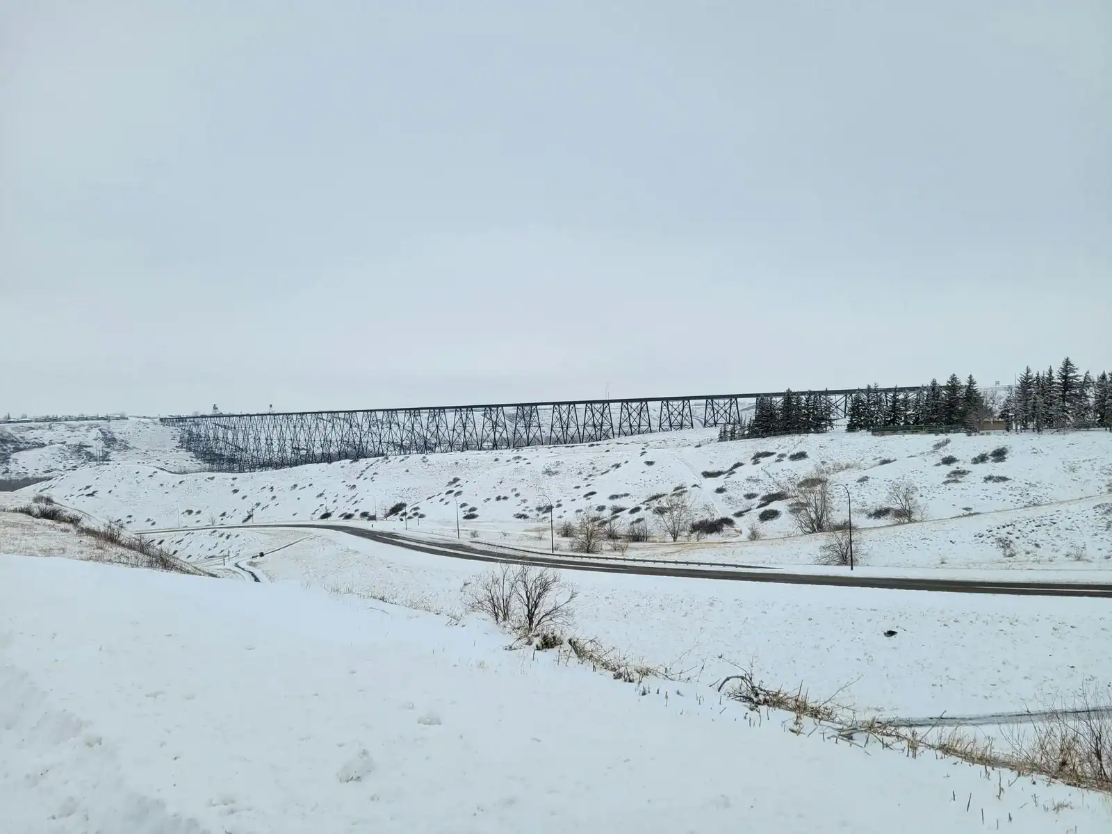 A tall steel railway bridge spans a snowy valley under a gray sky, with a road curving through the snow-covered landscape in the foreground. Sparse trees line the right side of the scene, much like how a skilled Lethbridge web design company shapes seamless digital paths through creative landscapes. - Clio Websites | Top Canadian Web Design Company