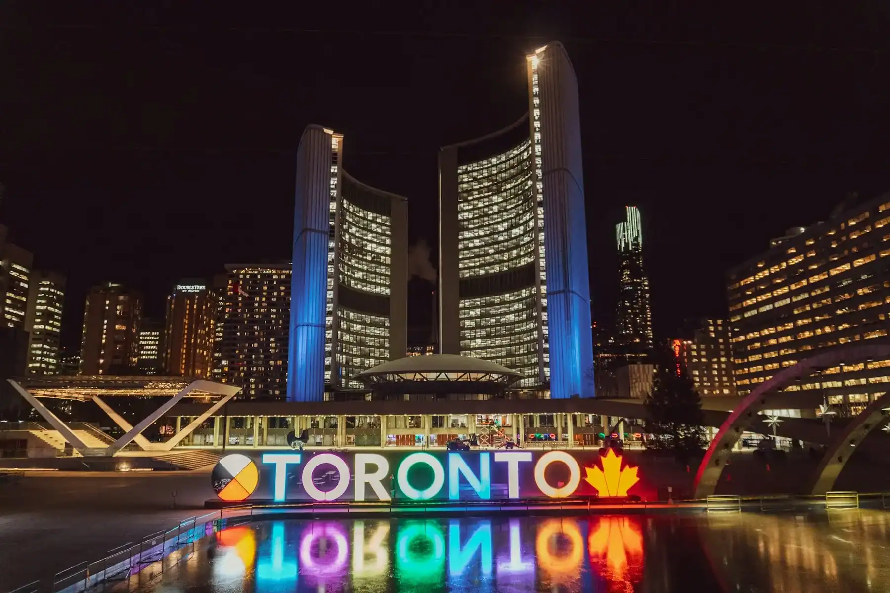 Toronto’s illuminated sign with colorful lights is in front of City Hall at night, reflected in a pool. Modern high-rise buildings surround the scene, and a red maple leaf symbolizes Canada—a vibrant setting that inspires creativity for any Ontario web design company. - Clio Websites | Top Canadian Web Design Company