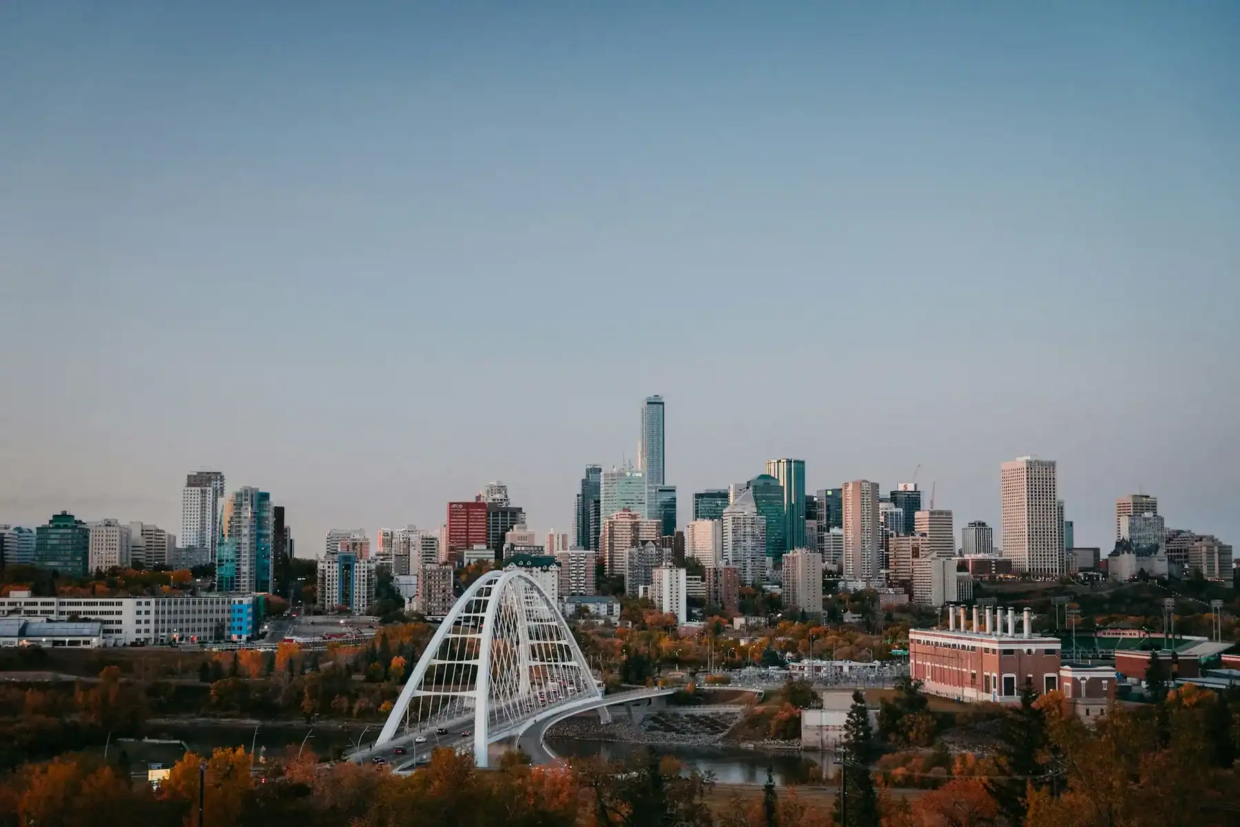 A city skyline with modern high-rise buildings, a white arched bridge, and a river surrounded by autumn trees under a clear blue sky—much like the inspiring backdrop that motivates creativity at any leading Saskatoon web design company. - Clio Websites | Top Canadian Web Design Company