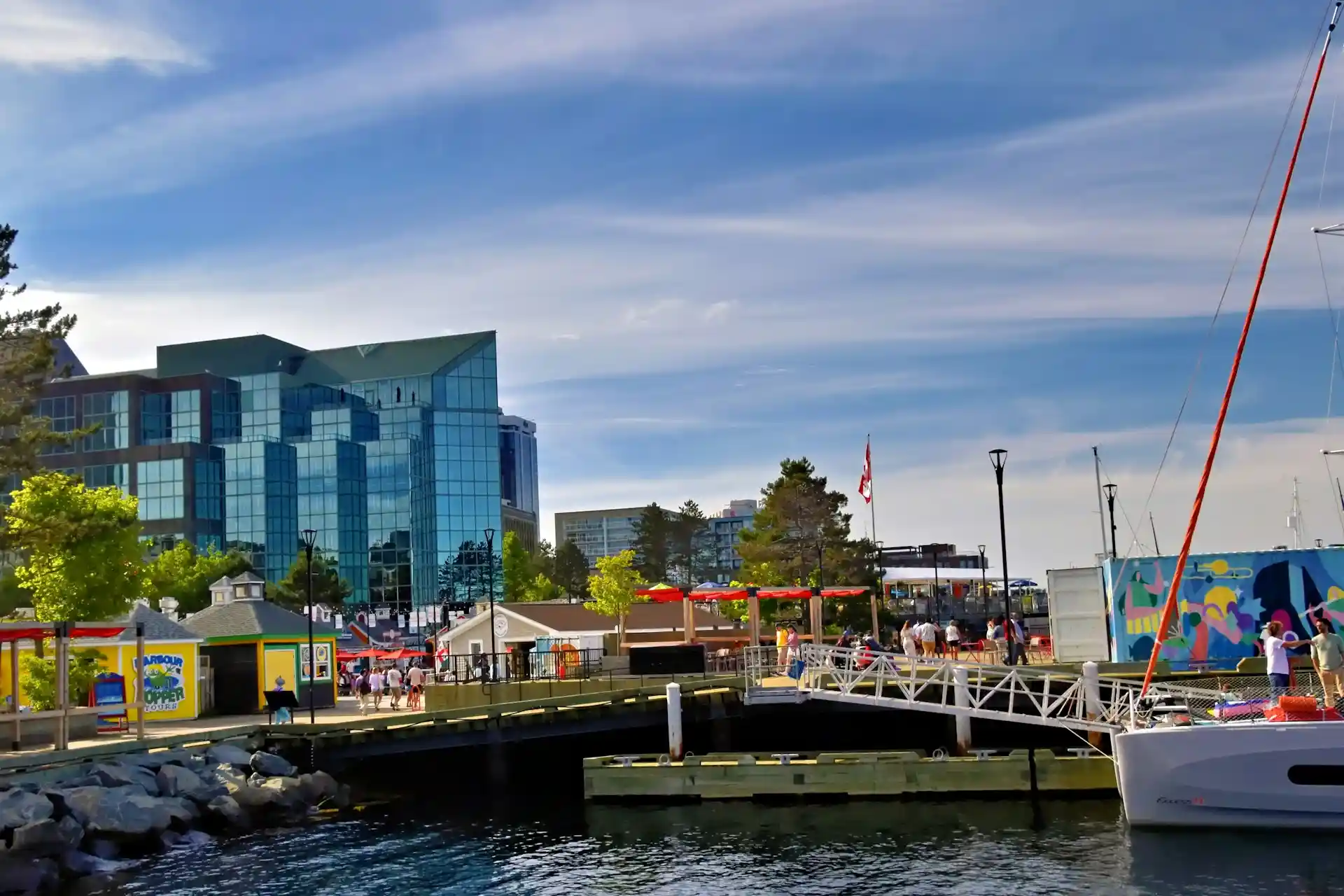 A waterfront scene with modern glass buildings, people walking along the pier, boats docked, colorful tents, trees, and a blue sky with wispy clouds—an inspiring setting reminiscent of creativity and innovation found in Saskatoon web design. - Clio Websites | Top Canadian Web Design Company