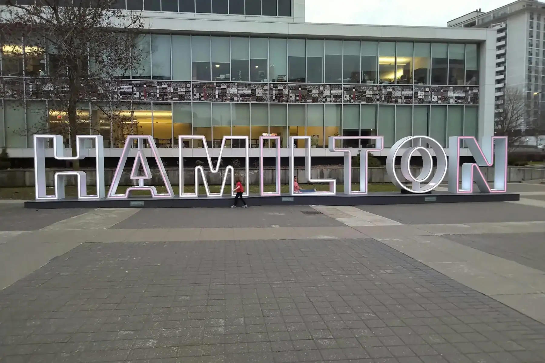 Large white letters spelling “HAMILTON” stand outside a modern glass building, representing the city’s vibrant innovation scene. A person in red stands between the letters “I” and “L.” The area is paved with tiles, surrounded by trees and city buildings, reflecting a setting where creative industries—like Hamilton web design companies—thrive. - Clio Websites | Top Canadian Web Design Company