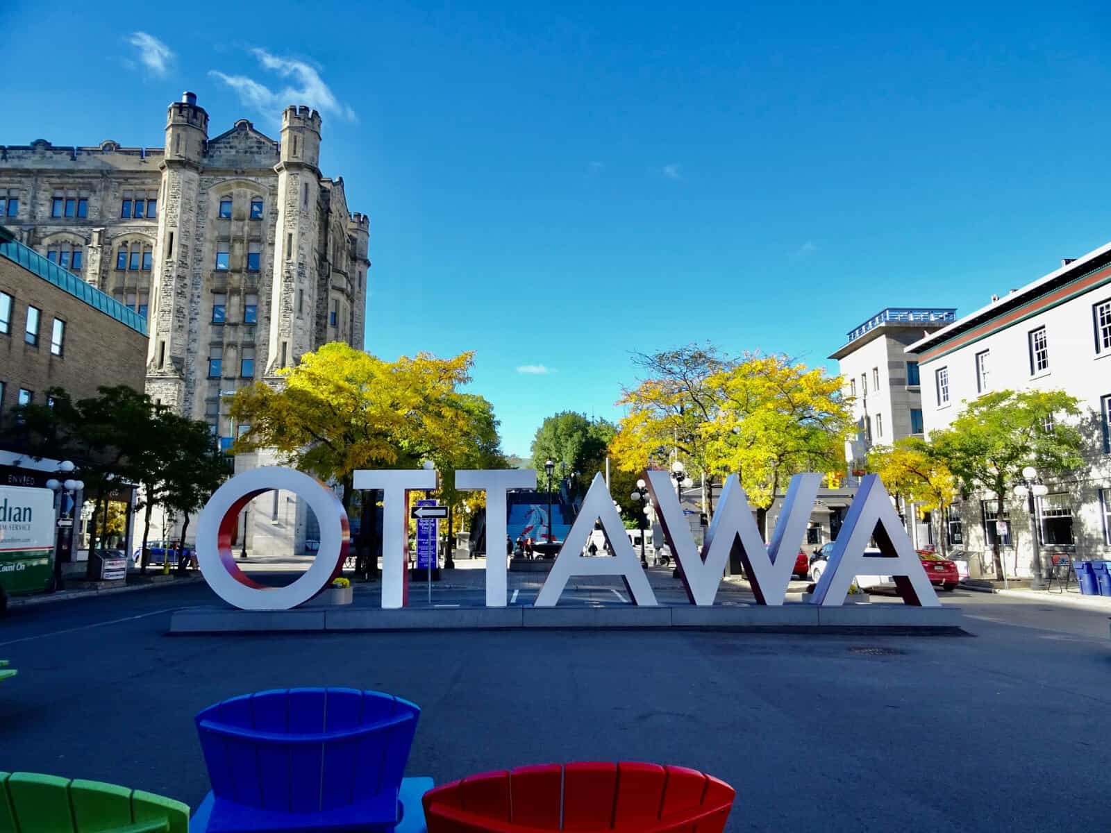 Large white letters spell “OTTAWA” on a city street, with historic buildings, trees with yellow leaves, and a bright blue sky in the background. Colorful chairs are visible in the foreground, creating a vibrant scene reminiscent of the creative flair you’d expect from a top Saskatoon web design company or experts in WordPress development Saskatoon. - Clio Websites | Top Canadian Web Design Company