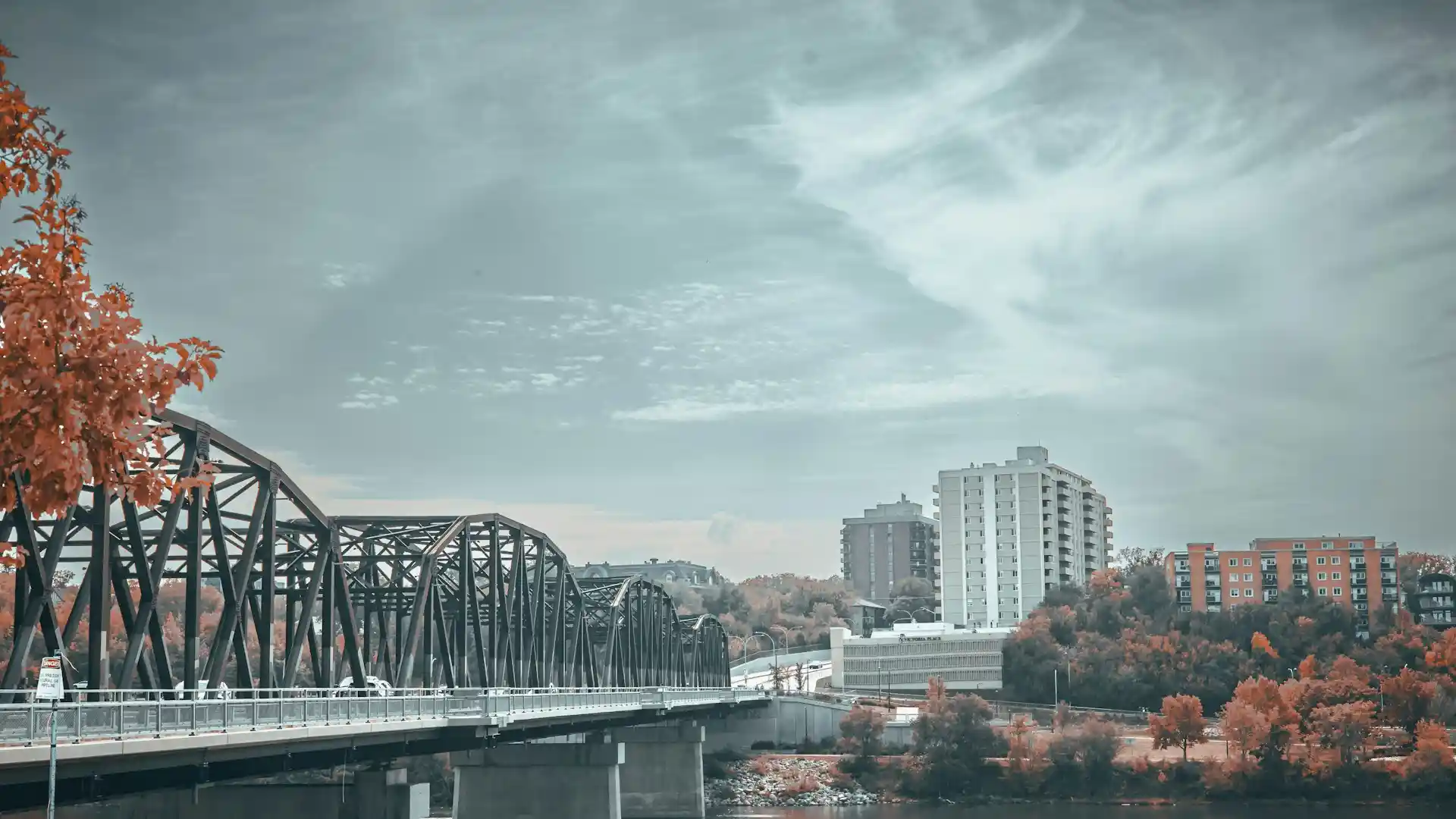 A steel truss bridge crosses a river with cars driving on it. Red-orange trees line the riverbank, and several tall apartment buildings stand in the background under a cloudy sky—a scene that captures the vibrant atmosphere of a city known for innovation, such as Saskatoon web design. - Clio Websites | Top Canadian Web Design Company