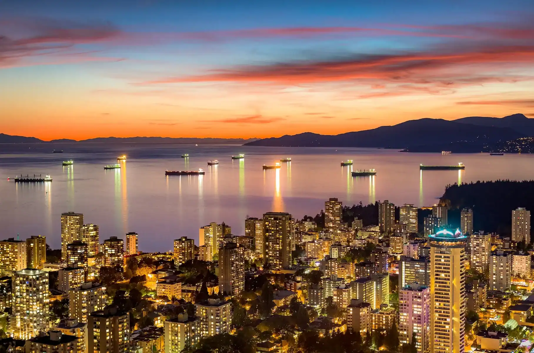 City skyline at dusk with illuminated buildings, calm water reflecting city lights, and several ships anchored in the bay under a vibrant orange and blue sunset sky with distant mountains—a captivating scene reminiscent of the innovative energy that drives a leading Saskatoon web design company, blending creativity and functionality much like the colors of this stunning urban horizon. - Clio Websites | Top Canadian Web Design Company
