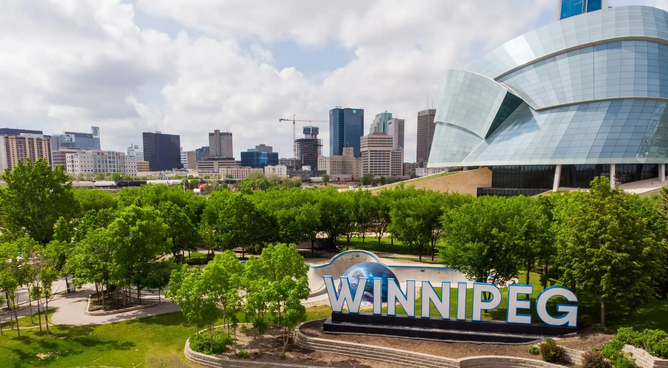 A large “Winnipeg” sign stands in front of trees, with modern city buildings and the Canadian Museum for Human Rights visible in the background under a partly cloudy sky, much like how Saskatoon website design blends urban innovation with natural beauty—a hallmark of any leading Saskatoon web design company. - Clio Websites | Top Canadian Web Design Company