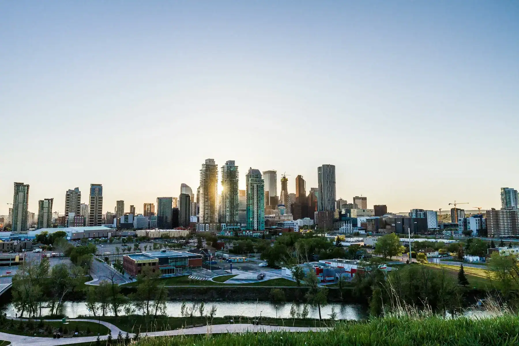 City skyline at sunrise or sunset with modern high-rise buildings, a river in the foreground, and green spaces, all under a clear sky—an inspiring scene that reflects the innovation and creativity of a leading Saskatoon web design company. - Clio Websites | Top Canadian Web Design Company