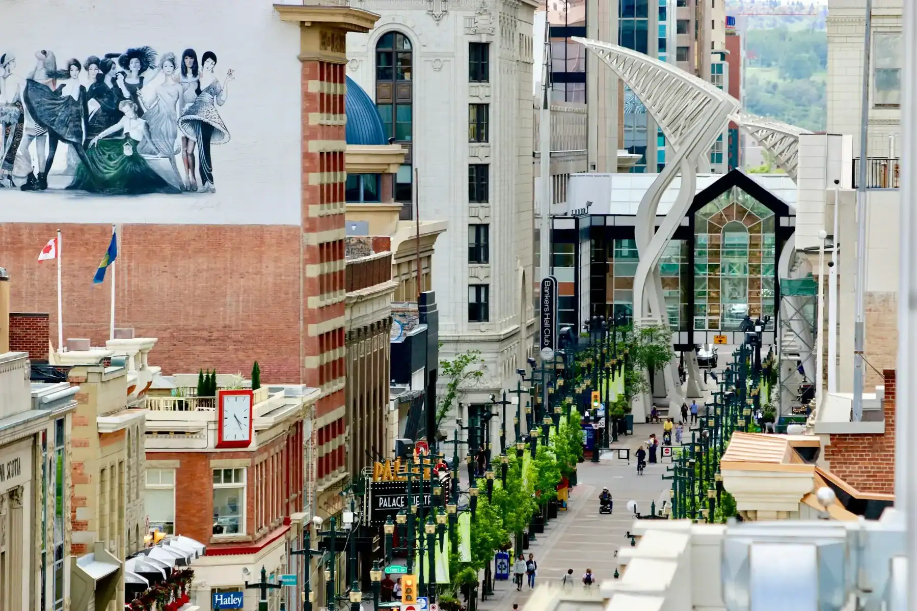 A busy pedestrian street with shops, greenery, and people. Large mural of dancers on a building, modern sculpture, and a glass-covered entrance are visible. The Canadian flag and cityscape in the background reflect the dynamic spirit of Saskatoon—a city also known for its thriving creative scene, including Saskatoon web design and WordPress development services. - Clio Websites | Top Canadian Web Design Company