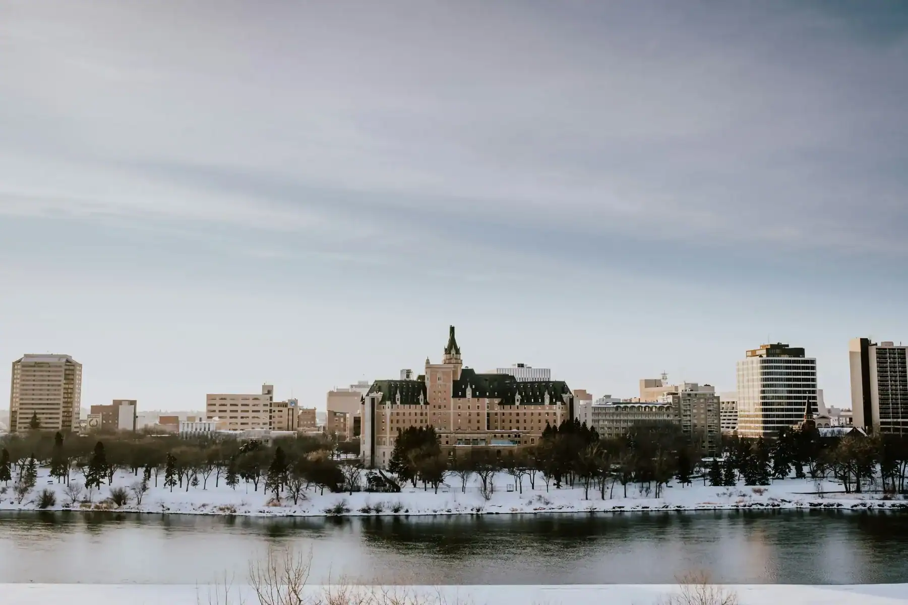 City skyline in winter, with snow-covered ground and trees along a riverbank, and a tall historic building at the center under a cloudy blue sky—a scene reminiscent of the inspiring creativity found at a leading Saskatoon web design company. - Clio Websites | Top Canadian Web Design Company