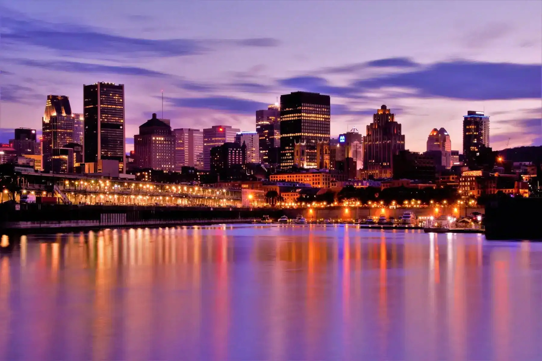 City skyline at dusk with tall buildings illuminated, reflecting on calm water below. The sky is purple and blue with scattered clouds, and city lights create a warm glow along the waterfront—an inspiring view reminiscent of creativity and innovation found at a leading Saskatoon web design company. - Clio Websites | Top Canadian Web Design Company